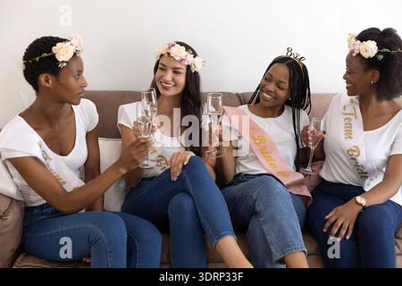 Verschiedene Freundinnen in Schärpen feiern Junggesellinnenabschied auf der Couch mit Sektflöten. Hochzeitsdusche, Feier, Stockfoto