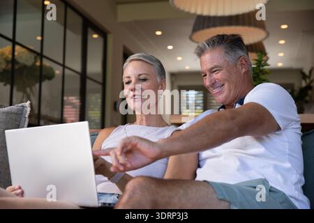 Ein älteres Paar sitzt auf der Couch im offenen Zimmer mit silbernem Laptop und zeigt auf den Bildschirm. Modern, gemütlich, modern, gemeinschaftlich Stockfoto