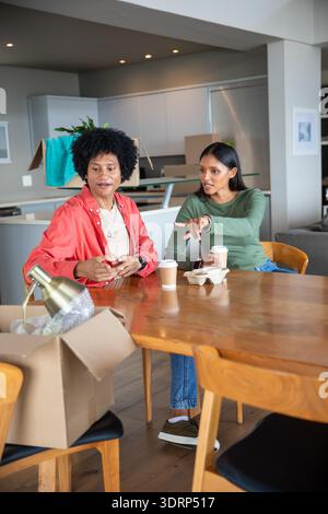 Afroamerikaner und indischer Paare packen Box in moderner Küche mit Messinglampe und Kaffeetassen aus. Umzug, Umzug, Zuhause, Einzug Stockfoto