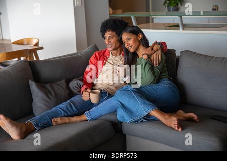 Paare sitzen auf einem Sofa im Wohnzimmer und halten hellbraune Keramikbecher und Smartphone. Gemütlichkeit, Intimität, Entspannung, Wohnkultur, Verachtung Stockfoto
