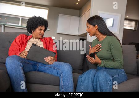 Afroamerikanisches und asiatisch-indisches Paar, das im Wohnzimmer mit Tablet und Smartphone auf einem Sofa sitzt. Familie, Kommunikation, modern, Stockfoto