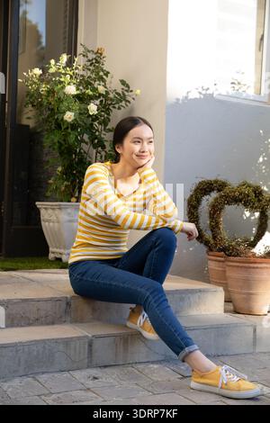 Asiatische Frau sitzt auf Steintreppen auf der Veranda und bewundert weiße Pflanzgefäße mit Rosen und Terrakottatöpfen. Ruhe, draußen, einladend, Stockfoto