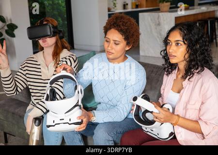 Verschiedene Freundinnen sitzen auf einem grauen Sofa und erkunden Virtual-Reality-Headsets in einem modernen Zuhause. Technologie, Entspannung, kollabora Stockfoto