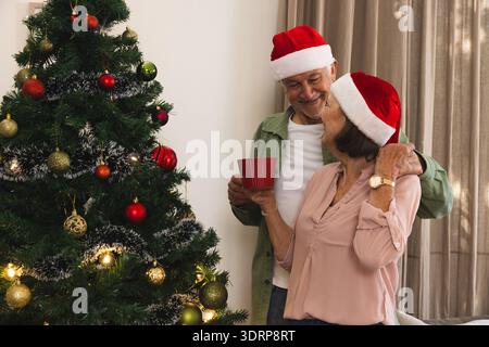 Seniorenpaar mit Weihnachtsmützen, die zu Hause am Weihnachtsbaum stehen, roten Becher halten, Kopierraum. Festlich, feierlich, warm, A Stockfoto