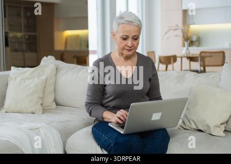 Selbstbewusst stilvoll glückliche Seniorin mittleren Alters, die zu Hause ein Notebook nutzt. Stilvolle ältere Großmutter aus den 60er Jahren, die auf der Couch sitzt Stockfoto