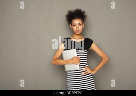 Die junge Frau steht vor einer grauen Wand und hält einen Laptop in einem schlichten Schwarz-weiß-Kleid Stockfoto