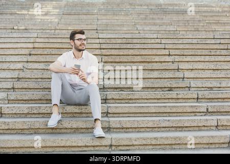 Der Mann sitzt tagsüber auf einer Treppe mit Telefon in einem öffentlichen Raum Stockfoto