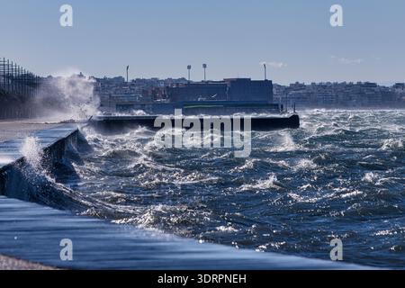 Mächtige Wellen krachen gegen die Uferpromenade in Thessaloniki und erzeugen dramatische Spritzer entlang der gekrümmten Küste. Ein Mensch und ein Hund gehen Stockfoto