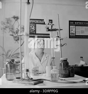 Historisches Schwarzweiß-Foto aus den 1960er Jahren, das eine Laborarbeiterin in der Öl- und Fettfabrik Slawjansk in der ukrainischen SSR zeigt. Mit weißem Mantel und Kappe sitzt sie an einem Schreibtisch mit chemischen Gläsern, Flaschen und Prüfgeräten für die Qualitätskontrolle. Der Hintergrund zeigt authentische sowjetische Arbeitsplatzpropaganda, einschließlich eines gewerkschaftsvorstands mit einem Hammer- und Sichelsymbol und einem Poster mit der Aufschrift „für Qualität“. Dieses Archivbild zeigt die Industriewissenschaft und die Frauenarbeit in der Region Donezk. Stockfoto