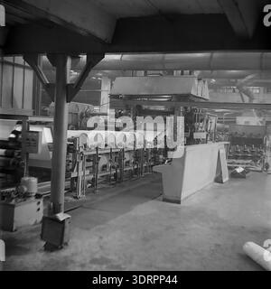 Das historische Schwarzweißfoto aus den 1960er Jahren zeigt das industrielle Innere einer Werkstatt in der Öl- und Fettfabrik Slawjansk in der ukrainischen SSR, UdSSR. Das Bild zeigt eine riesige Produktionslinie mit breiten Rollen aus weißem Verpackungsmaterial, die durch komplexe mechanische Rollen und schwere Maschinen transportiert werden. Zwei Arbeiter sind an einer Kontrollstation im Hintergrund der geräumigen Fabrikhalle zu sehen. Dieses authentische Archivfoto zeigt sowjetische Ingenieurskunst, industrielle Macht und die optimistische Atmosphäre im friedlichen Donbass vor dem Krieg. Stockfoto