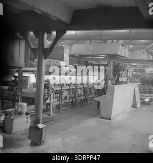 Das historische Schwarzweißfoto aus den 1960er Jahren zeigt das industrielle Innere einer Werkstatt in der Öl- und Fettfabrik Slawjansk in der ukrainischen SSR, UdSSR. Das Bild zeigt eine riesige Produktionslinie mit breiten Rollen aus weißem Verpackungsmaterial, die durch komplexe mechanische Rollen und schwere Maschinen transportiert werden. Zwei Arbeiter sind an einer Kontrollstation im Hintergrund der geräumigen Fabrikhalle zu sehen. Dieses authentische Archivfoto zeigt sowjetische Ingenieurskunst, industrielle Macht und die optimistische Atmosphäre im friedlichen Donbass vor dem Krieg. Stockfoto