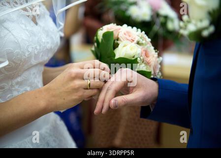 Aus nächster Nähe eine junge Braut, die während einer Winterzeremonie einen goldenen Ehering auf die Hand des Bräutigams legt. Weiche weiße Kleiddetails und Pastellbouqu Stockfoto