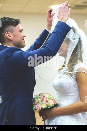 Der junge Bräutigam hebt den Schleier der Braut während einer Indoor-Winterhochzeit. Weiches weißes Kleid, Pastellstrauß und emotionale Ausdrucksformen Stockfoto