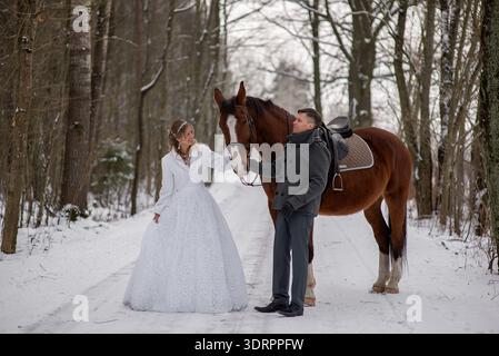 Frisch vermählte stehen auf einer verschneiten Waldstraße und halten ein ruhiges Pferd, das rustikalen Landschaftscharakter mit eleganter Winterhochzeitatmosphäre verbindet. Stockfoto