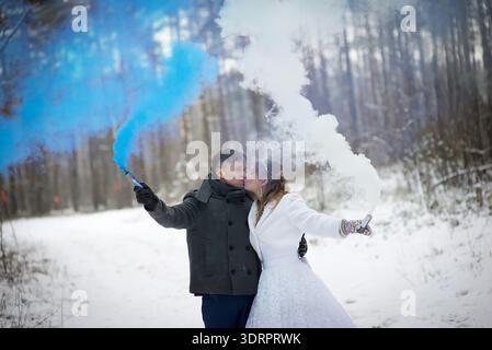 Horizontales Foto einer Braut und eines Bräutigams, die sich in einem verschneiten Wald küssen, während sie blaue und weiße Rauchfackeln halten, um einen intimen und romantischen Gewinn zu erzielen Stockfoto