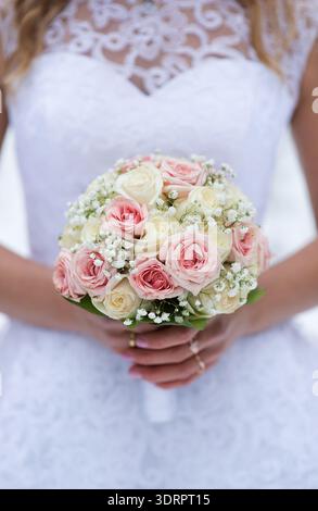 Vertikale Nahaufnahme eines Brautstraußes mit rosa und cremefarbenen Rosen und zarten weißen Blumen, gehalten von einer jungen erwachsenen Braut in einem weißen Spitzenkleid, Stockfoto