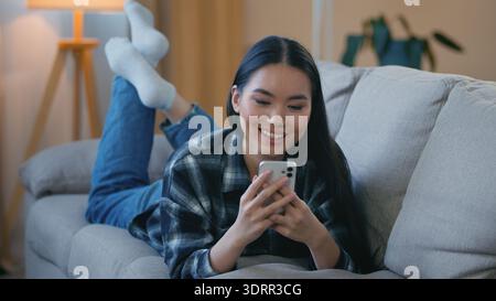 Lächelnde koreanische Frau liegend Sofa zu Hause entspannend mit Smartphone Asiatische weibliche Frau genießt Chat Tippen App Handy anschauen virales Video spielen Stockfoto