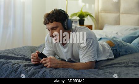 Fröhlicher kaukasischer Mann liegend Bett zu Hause mit Kopfhörern mit Smartphone singend Tanz glücklicher männlicher Kerl genießt Musik Song Melodie Audio Elektronik Stockfoto