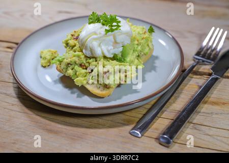 Sandwich mit Guacamole und einem pochierten Ei auf einem Teller und einem rustikalen Holztisch, köstlicher Frühstücks- oder Mittagssnack, ausgewählter Fokus, enge Tiefe des fleisches Stockfoto
