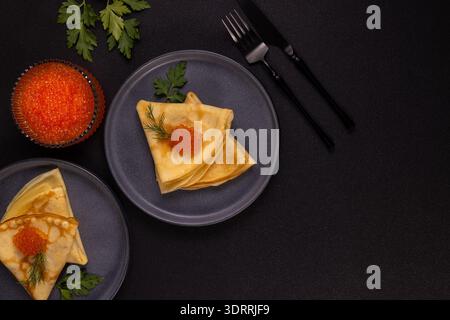 Zarte Blini-Pfannkuchen, die Teller mit leuchtendem rotem Lachskaviar füllen, perfekt auf dunklem Hintergrund neben einer vollen Schüssel Gourmet-Meeresfrüchten angeordnet, Stockfoto