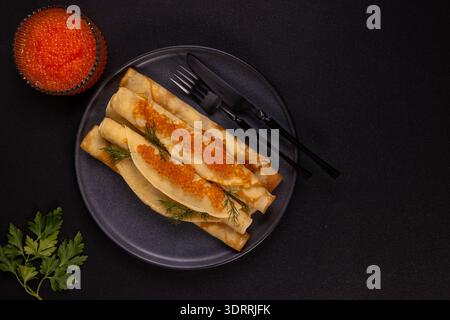 Dünne Pfannkuchen, gerollt mit hellrotem Lachskaviar und frischem Dill auf einem dunklen Servierteller mit einer Schüssel Reh und einem Zweig Petersilie, die einen Gourmets schaffen Stockfoto