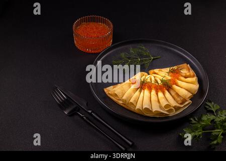 Dünne Blini-Pfannkuchen, gefächert auf einem dunklen Teller, gekrönt mit leuchtend rotem Kaviar und Dill, mit einer kleinen Schüssel mit Reh und schwarzem Besteck für einen eleganten Gourmets Stockfoto
