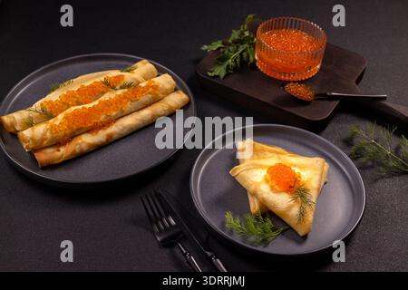 Zarte, gerollte und gefaltete Kreppchen, großzügig belegt mit leuchtendem rotem Kaviar und aromatischem Dill, angeordnet auf dunklen Tellern für einen eleganten Gourmet-Vorgeschmack Stockfoto