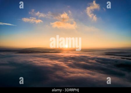 Aus der Vogelperspektive geht die Sonne über der Wolkendecke auf und wirft warmes goldenes Licht über die nebelige Landschaft. Ein heiterer Himmel mit sanften Pastelltönen, während die Gipfel weit entfernter Berge aus dem Nebel unter sich auftauchen. Stockfoto