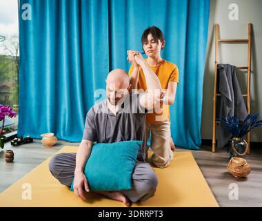Masseurin, die thai-Yoga-Massage macht. Frau Therapeutin, die traditionelle Massagebehandlung für männlichen Patienten in Innenräumen durchführt. Glatzköpfiger Kunde sitzt auf der Matte, meditiert und genießt eine Handmassage in ruhiger Atmosphäre. Stockfoto