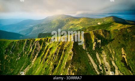 Atemberaubender Blick aus der Luft auf grüne, zerklüftete Berggipfel unter dramatischem Himmel. Die Szene fängt den Kontrast zwischen lebhafter Landschaft und stimmungsvollen Wolken ein, wobei Sonnenlicht durchbricht. Karpaten, Chornogora Stockfoto
