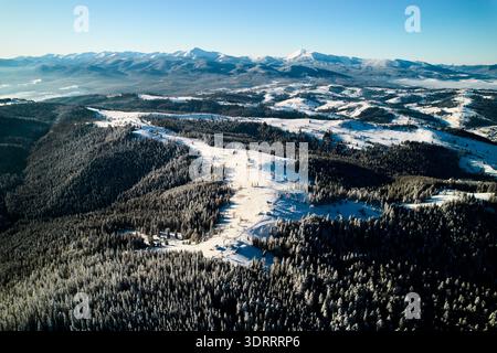 Blick aus der Vogelperspektive auf das schneebedeckte Tal mit kurviger Straße, die durch ein kleines Dorf führt. Die Landschaft bietet sanfte Hügel, dichte Wälder und ferne Berge unter klarem blauen Himmel mit weichem Nebel. Stockfoto