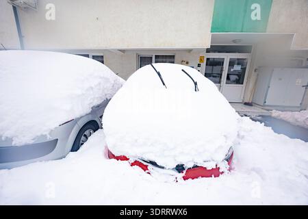 Rotes Auto, das vollständig unter einer dicken Neuschneeschicht mit Scheibenwischern begraben wurde, die nach einem schweren Wintersturm in einem Innenhof in einem Wohnhaus hochgehoben wurden Stockfoto
