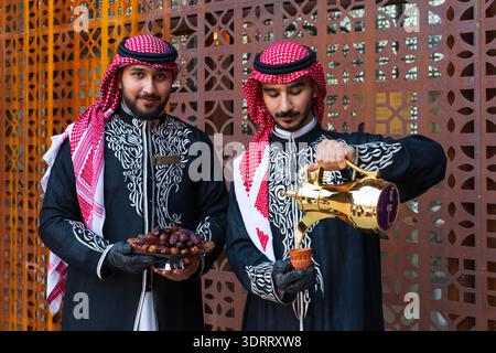 Zwei Männer in traditioneller saudischer Kleidung gießen Kaffee und servieren Datteln auf einer Platte in der Altstadt von Alula, Saudi-Arabien Stockfoto