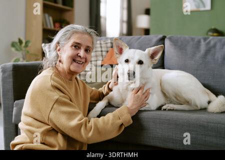 Porträt einer älteren kaukasischen Frau, die lächelt, während der Hund auf dem Sofa streichelt Stockfoto