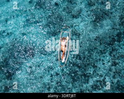 Aus der Vogelperspektive einer Frau, die sich in der Sonne auf einem transparenten Kajak sonnt, auf dem Zyanmeer, Anse Reunion, La Digue, Seychellen. Stockfoto