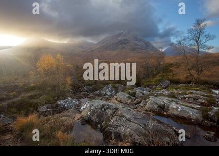 Sonnenlicht bricht durch Wolken über felsigen Bächen und beleuchtet Herbstbäume vor der Kulisse von Berggipfeln in den schottischen Highlands. Stockfoto