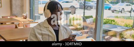 Ein stilvoller junger Mann mit Kopfhörern lächelt, während er draußen sein Handy überprüft. Stockfoto