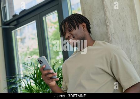 Ein hübscher junger Mann lächelt, als er sein Handy überprüft, umgeben von Grün und Licht. Stockfoto
