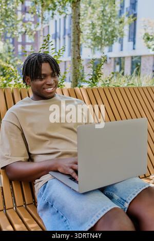 Ein hübscher junger Mann konzentriert sich auf seinen Laptop und lächelt, während er die ruhige Parklandschaft genießt. Stockfoto