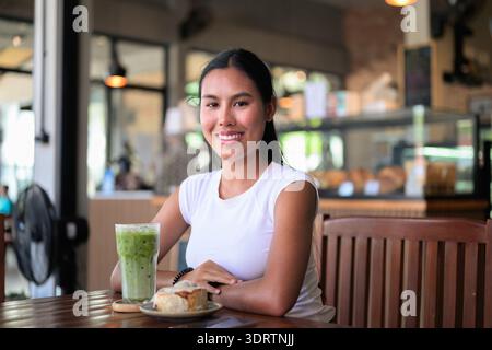 Lächelnde asiatische Frau trinkt Iced Matcha Latte im Café mit Nachtisch Stockfoto