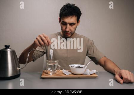 Studio-Porträt eines jungen Mannes, der warmen Tee zubereitete, einen tropfenden Teebeutel über einem mit heißem Wasser gefüllten Glasbecher hielt, um ein tröstliches Morgenritual zu genießen Stockfoto