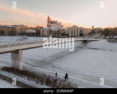 Aus der Vogelperspektive auf den schneebedeckten Neris, der unter der Mindaugas-Brücke fließt und sanftes Sonnenlicht in der Nähe der St. Peter- und St. Paul-Kirche in Vilnius reflektiert. Stockfoto
