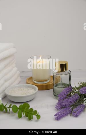 Minimaler Wellness-Hintergrund mit Kopierraum: Handtücher, beleuchtete Kerze auf Bambusuntersetzer, Badesalz, Lavendel, Eukalyptus und eine Glasduftflasche auf mA Stockfoto