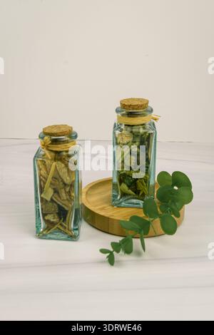 Natürliches Wellness-Stillleben: Zwei Gläser mit Korkdeckel gefüllt mit getrockneten Pflanzenstoffen und Kräutern auf Bambustablett, Eukalyptuszweig, Marmorhintergrund Stockfoto