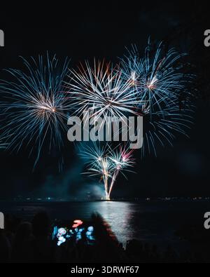 Blick auf das pulsierende Feuerwerk, das über dem dunklen Wasser explodiert und sich in der ruhigen Oberfläche spiegelt, während Menschenmassen vom Ufer, Neuchâtel, Neuchâtel, Switzer, beobachten Stockfoto