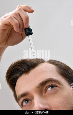 Nahaufnahme eines Mannes, der nach oben blickt, während er flüssiges Serum mit Tropfer auf das Haar auf hellgrauem Hintergrund in minimaler Studioeinstellung aufträgt Stockfoto