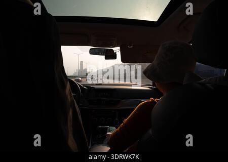 Innenansicht vom Auto aus mit Fahrer und Beifahrer auf der Autobahn mit der Skyline der Stadt im Hintergrund bei Sonnenuntergang Stockfoto