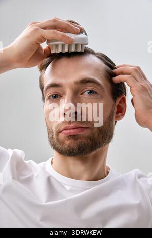 Nahaufnahme eines erwachsenen bärtigen Mannes in weißem T-Shirt, der mit einer Hand Kopfbürste an die Haare drückt, während er mit der anderen den Tempel berührt Stockfoto