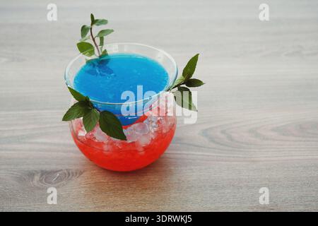 Helles blaues und rotes Getränk, serviert in einem runden Glas auf einem Holztisch mit Eiswürfeln Stockfoto