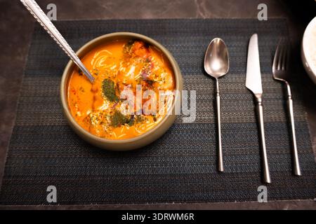 Indische Curry-Suppe in Keramikschale mit Löffel und Besteck auf einem Tischset mit dunkler Struktur Stockfoto